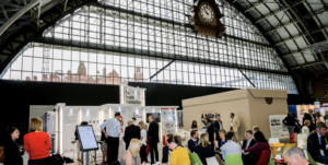 A photo of people in a tall building, chatting in small groups and dressed smartly. Behind them is a giant shoebox with an open door on one side, large enough to walk into.